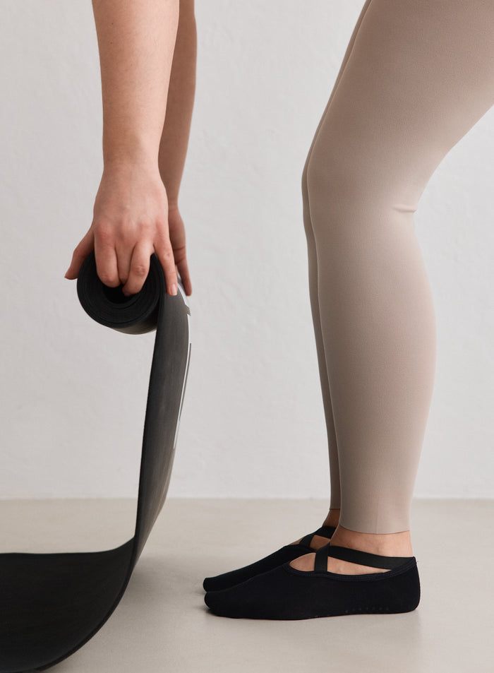 Henkilö yllään mustat aim'n Pilates Socks -sukat ja beiget trikoot, pitelemässä rullattua mustaa jumppamattoa.