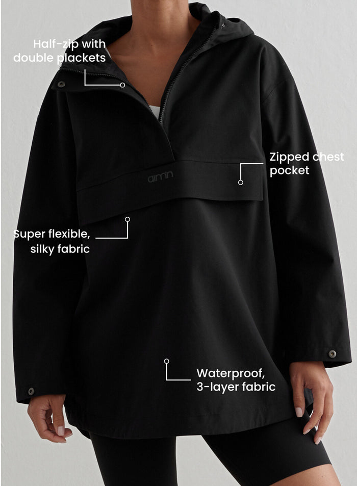 Lähikuva henkilöstä, jolla on päällään musta aim'n Waterproof Oversized Anorak. Kuvassa on infotekstejä, jotka korostavat toiminnallisia yksityiskohtia, kuten puolivetoketjua tuplanapituslistalla, vetoketjullista rintataskua ja vedenpitävää 3-kerroksista kangasta.