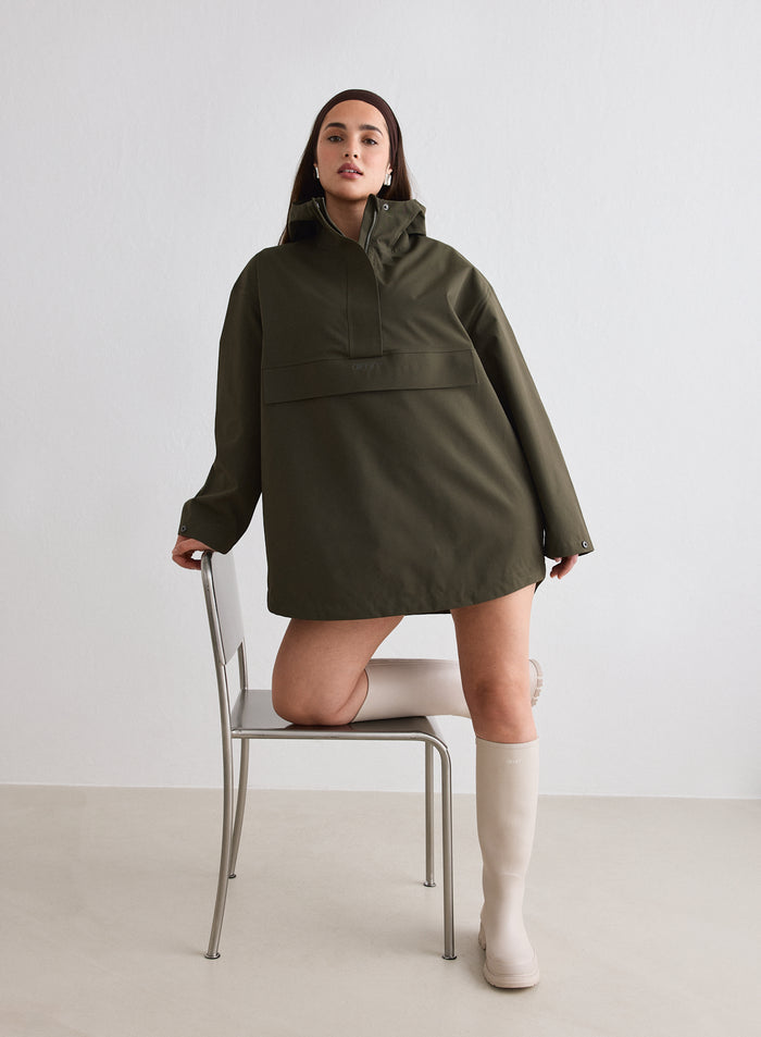 Malli yllään aim'n Khaki Waterproof Oversized Anorak ja korkeat beiget saappaat, poseeraa yksi polvi tuolilla [M-174cm]