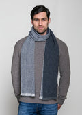 Man wearing a gray sweater and blue jeans with a large gray scarf draped over his shoulders against a plain background.