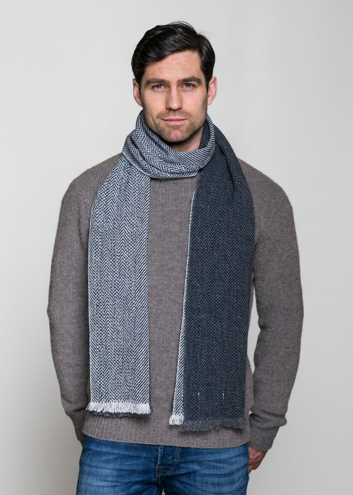 Man wearing a gray sweater and blue jeans with a large gray scarf draped over his shoulders against a plain background.