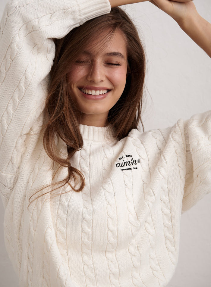 Hymyilevä nainen yllään luonnonvalkoinen aim'n Club Cable Knit Sweater logoyksityiskohdalla. [M-174cm]